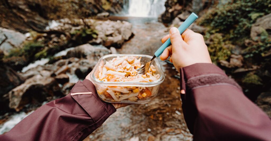 キャンプ場で家族が昼食を囲んで笑顔で過ごす風景。テーブルに並ぶ彩り豊かな料理と子どもたちの楽しそうな表情が印象的。